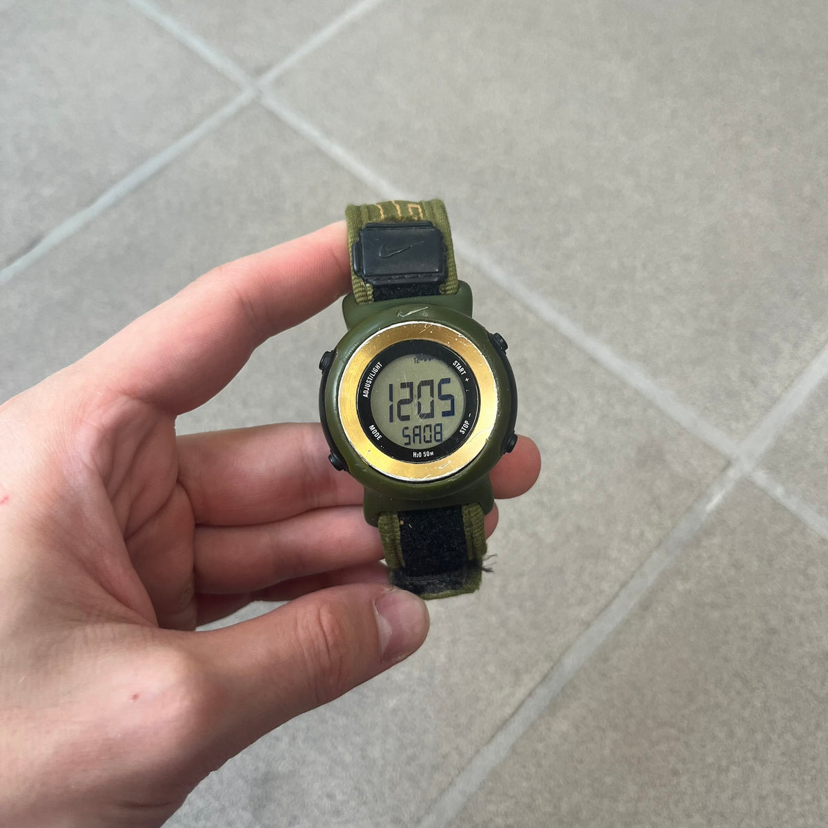 Nike 2000's Gorge Sport WK0013 Digital Watch is a vintage digital watch featuring a green circular face framed by a gold ring, water resistant up to 50m. It's fitted with a green canvas strap accented with orange line patterns and a black leather tab bearing the Nike logo.