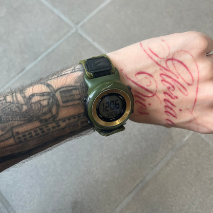Nike 2000's Gorge Sport WK0013 Digital Watch is a vintage digital watch featuring a green circular face framed by a gold ring, water resistant up to 50m. It's fitted with a green canvas strap accented with orange line patterns and a black leather tab bearing the Nike logo.