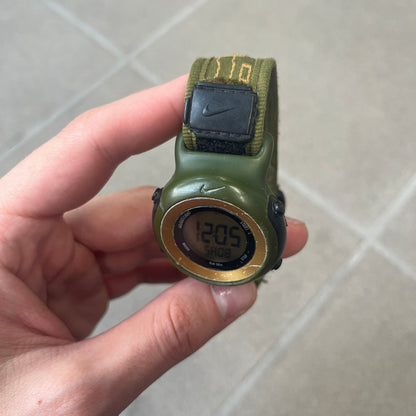 Nike 2000's Gorge Sport WK0013 Digital Watch is a vintage digital watch featuring a green circular face framed by a gold ring, water resistant up to 50m. It's fitted with a green canvas strap accented with orange line patterns and a black leather tab bearing the Nike logo.
