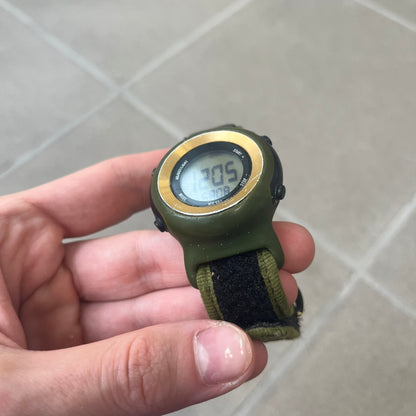 Nike 2000's Gorge Sport WK0013 Digital Watch is a vintage digital watch featuring a green circular face framed by a gold ring, water resistant up to 50m. It's fitted with a green canvas strap accented with orange line patterns and a black leather tab bearing the Nike logo.
