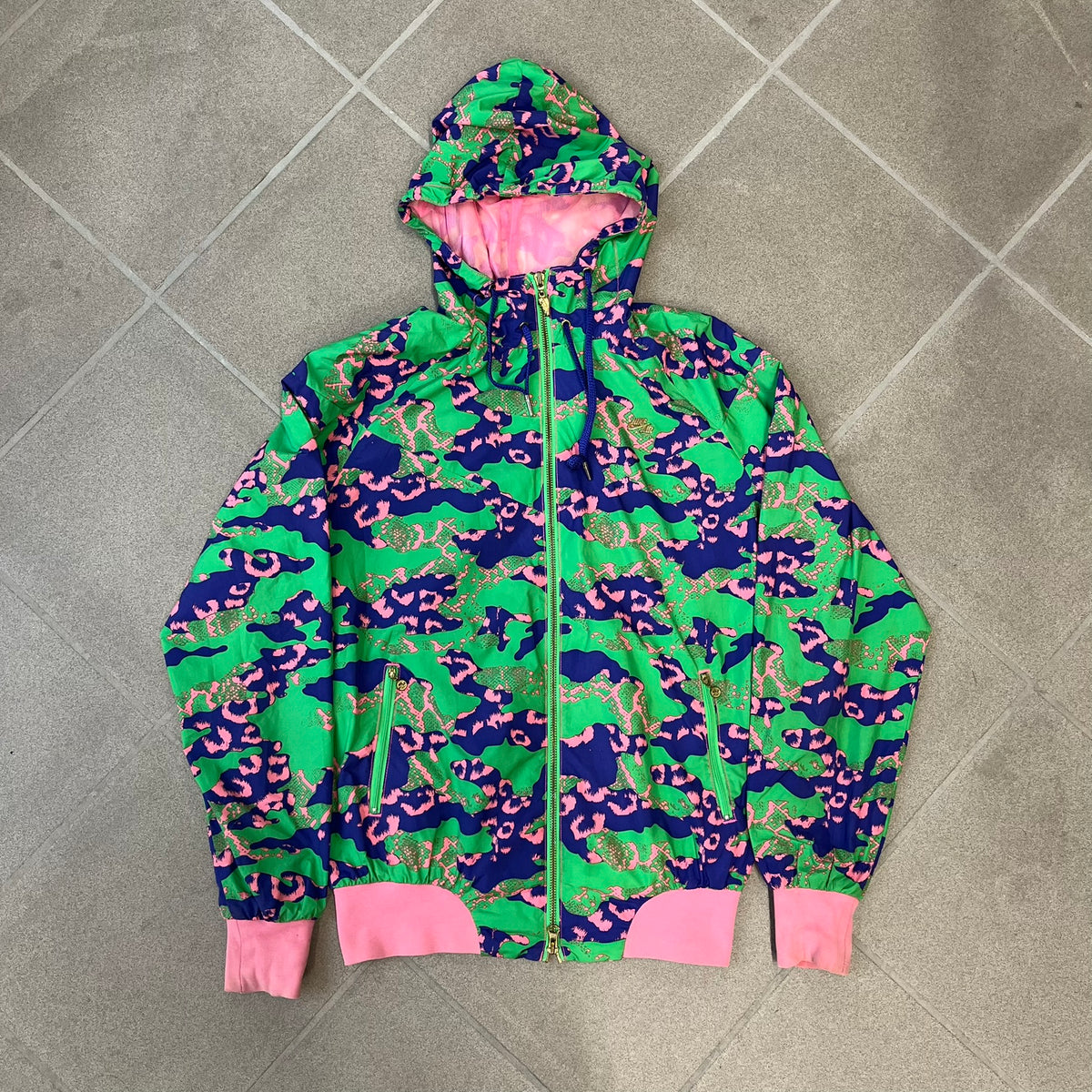 Nike 2000's Green & Pink Camo Windbreaker features an all-over camouflage pattern with a green base and pink and purple details. It has pink hems, a pink mesh lining, and a gold-tone 3D Nike shoe zipper pull, while the pocket zippers feature Nike Air logos. The chest has a gold-tone metal Nike Air logo.
