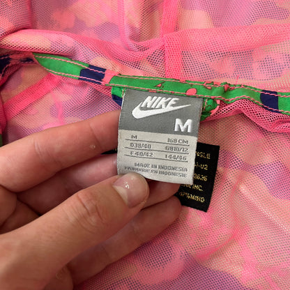 Nike 2000's Green & Pink Camo Windbreaker features an all-over camouflage pattern with a green base and pink and purple details. It has pink hems, a pink mesh lining, and a gold-tone 3D Nike shoe zipper pull, while the pocket zippers feature Nike Air logos. The chest has a gold-tone metal Nike Air logo.
