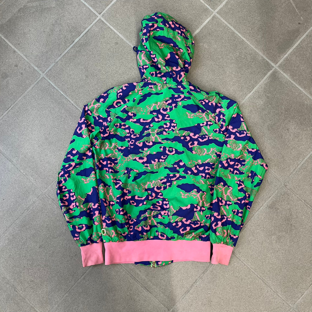 Nike 2000's Green & Pink Camo Windbreaker features an all-over camouflage pattern with a green base and pink and purple details. It has pink hems, a pink mesh lining, and a gold-tone 3D Nike shoe zipper pull, while the pocket zippers feature Nike Air logos. The chest has a gold-tone metal Nike Air logo.
