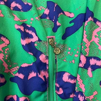 Nike 2000's Green & Pink Camo Windbreaker features an all-over camouflage pattern with a green base and pink and purple details. It has pink hems, a pink mesh lining, and a gold-tone 3D Nike shoe zipper pull, while the pocket zippers feature Nike Air logos. The chest has a gold-tone metal Nike Air logo.

