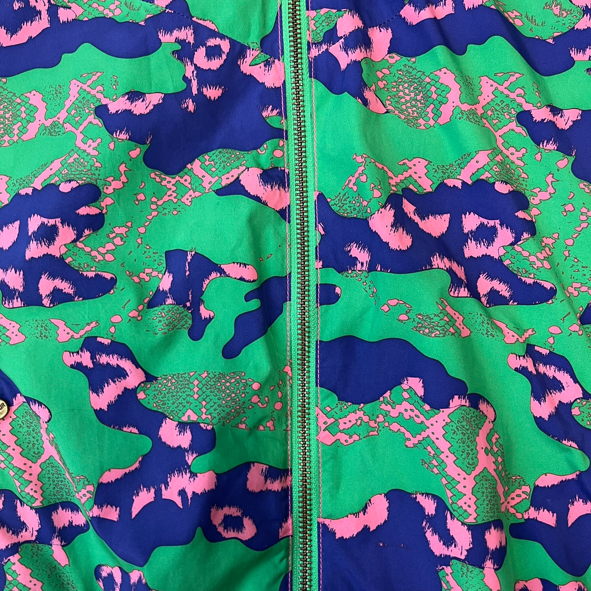 Nike 2000's Green & Pink Camo Windbreaker features an all-over camouflage pattern with a green base and pink and purple details. It has pink hems, a pink mesh lining, and a gold-tone 3D Nike shoe zipper pull, while the pocket zippers feature Nike Air logos. The chest has a gold-tone metal Nike Air logo.
