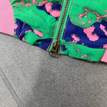 Nike 2000's Green & Pink Camo Windbreaker features an all-over camouflage pattern with a green base and pink and purple details. It has pink hems, a pink mesh lining, and a gold-tone 3D Nike shoe zipper pull, while the pocket zippers feature Nike Air logos. The chest has a gold-tone metal Nike Air logo.
