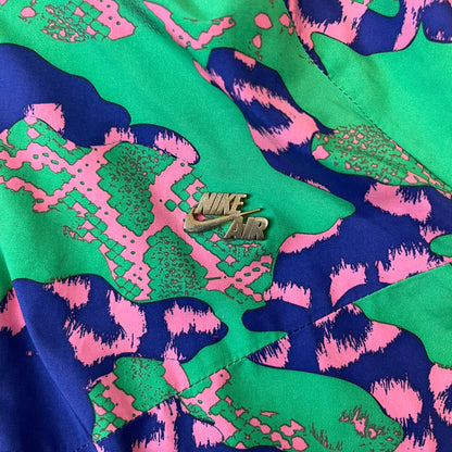Nike 2000's Green & Pink Camo Windbreaker features an all-over camouflage pattern with a green base and pink and purple details. It has pink hems, a pink mesh lining, and a gold-tone 3D Nike shoe zipper pull, while the pocket zippers feature Nike Air logos. The chest has a gold-tone metal Nike Air logo.
