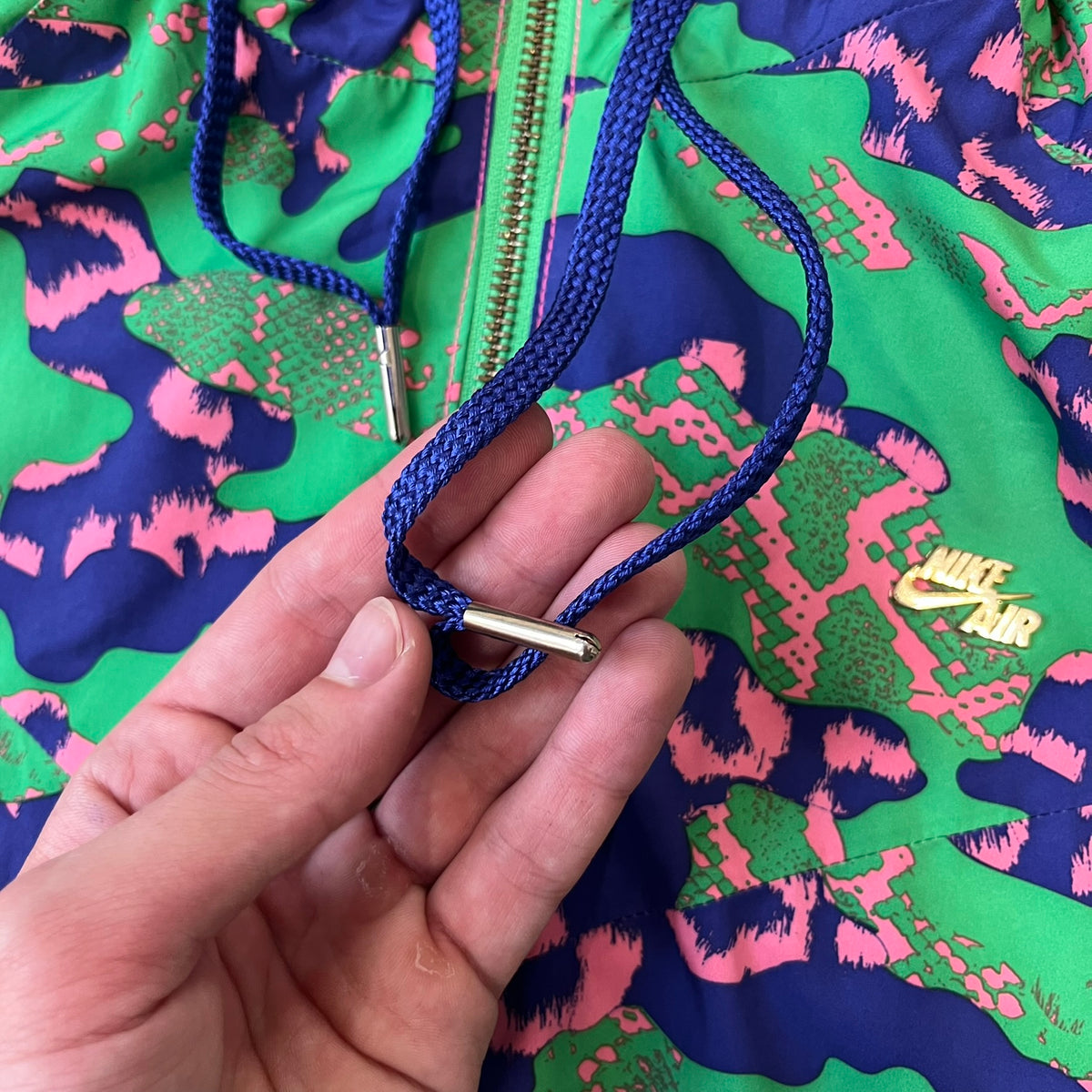 Nike 2000's Green & Pink Camo Windbreaker features an all-over camouflage pattern with a green base and pink and purple details. It has pink hems, a pink mesh lining, and a gold-tone 3D Nike shoe zipper pull, while the pocket zippers feature Nike Air logos. The chest has a gold-tone metal Nike Air logo.
