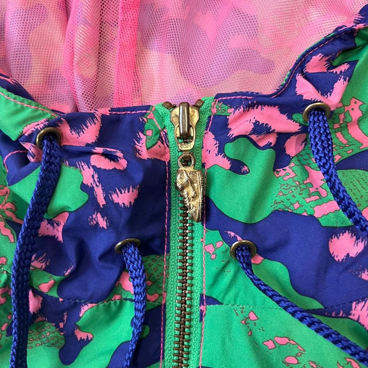 Nike 2000's Green & Pink Camo Windbreaker features an all-over camouflage pattern with a green base and pink and purple details. It has pink hems, a pink mesh lining, and a gold-tone 3D Nike shoe zipper pull, while the pocket zippers feature Nike Air logos. The chest has a gold-tone metal Nike Air logo.
