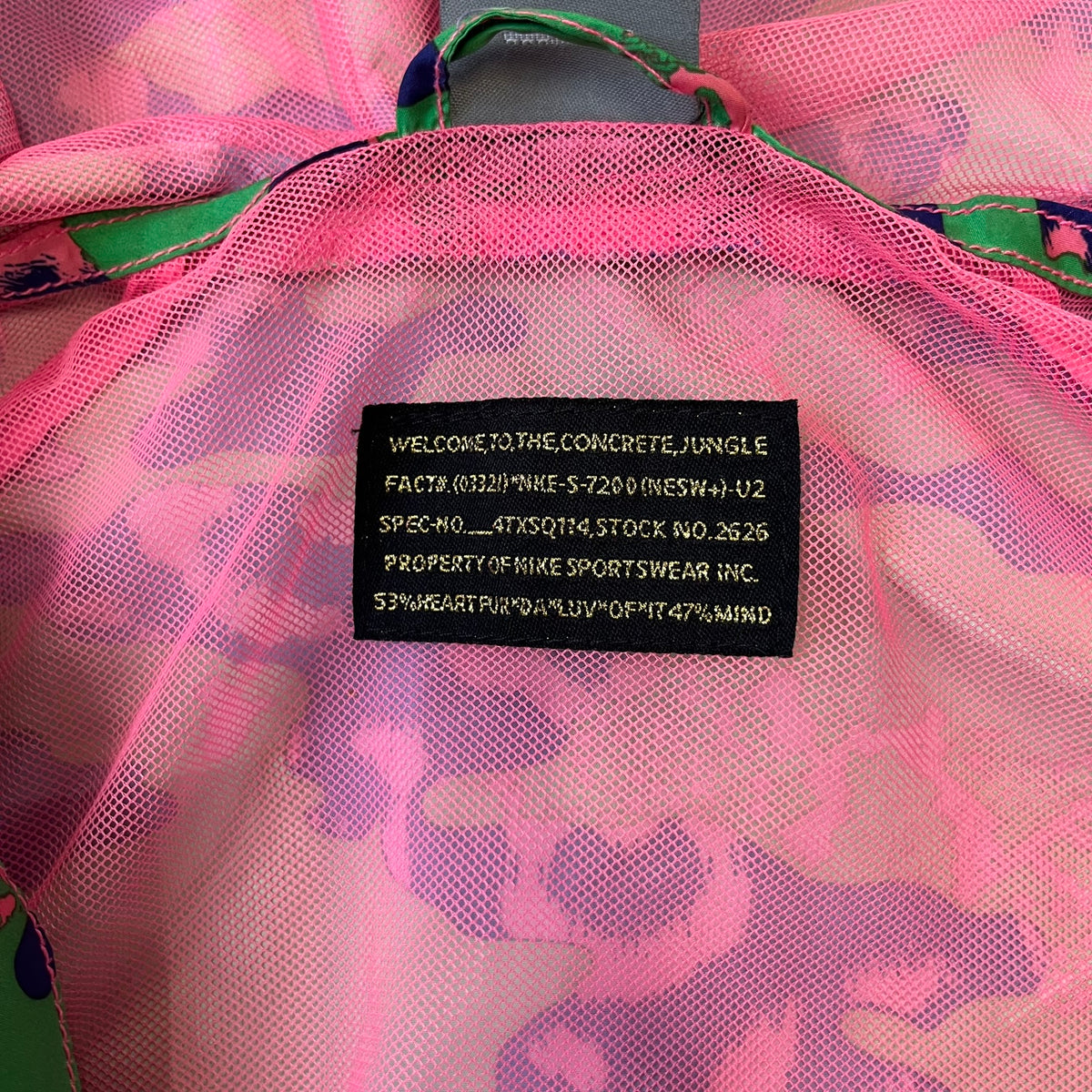 Nike 2000's Green & Pink Camo Windbreaker features an all-over camouflage pattern with a green base and pink and purple details. It has pink hems, a pink mesh lining, and a gold-tone 3D Nike shoe zipper pull, while the pocket zippers feature Nike Air logos. The chest has a gold-tone metal Nike Air logo.
