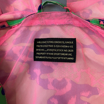 Nike 2000's Green & Pink Camo Windbreaker features an all-over camouflage pattern with a green base and pink and purple details. It has pink hems, a pink mesh lining, and a gold-tone 3D Nike shoe zipper pull, while the pocket zippers feature Nike Air logos. The chest has a gold-tone metal Nike Air logo.
