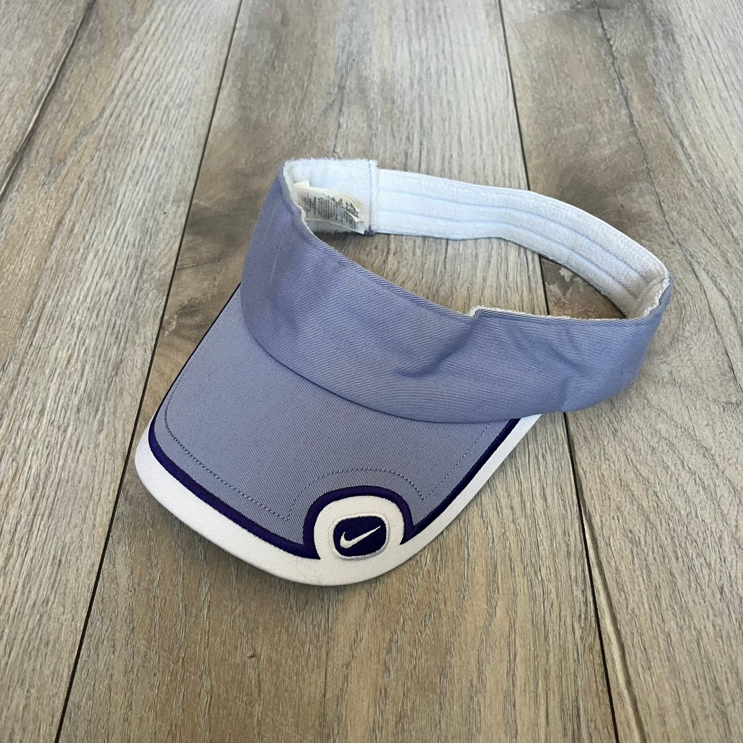Nike 2000s Violet &amp; White Tennis Visor is a youth-size visor cap featuring a violet crown with white trim running along the edge. The front includes a navy and white Nike logo, while the underside of the brim is finished in white. The visor is designed with a soft, comfortable headband-style strap for a secure fit.