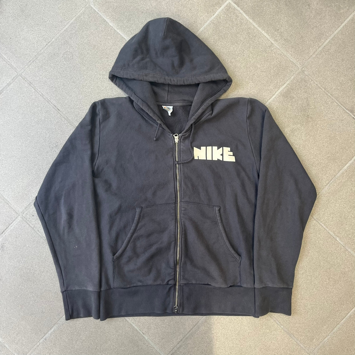 Nike 2010’s Big Logo Black Hoodie is a limited-edition black washed zip hoodie made in Portugal, with a white Nike logo on the chest and a larger one across the upper back, both non-standard designs. It has a standard zipper and a neck tag styled after Nike’s 1980s tags, and the whole piece is finished to look vintage.