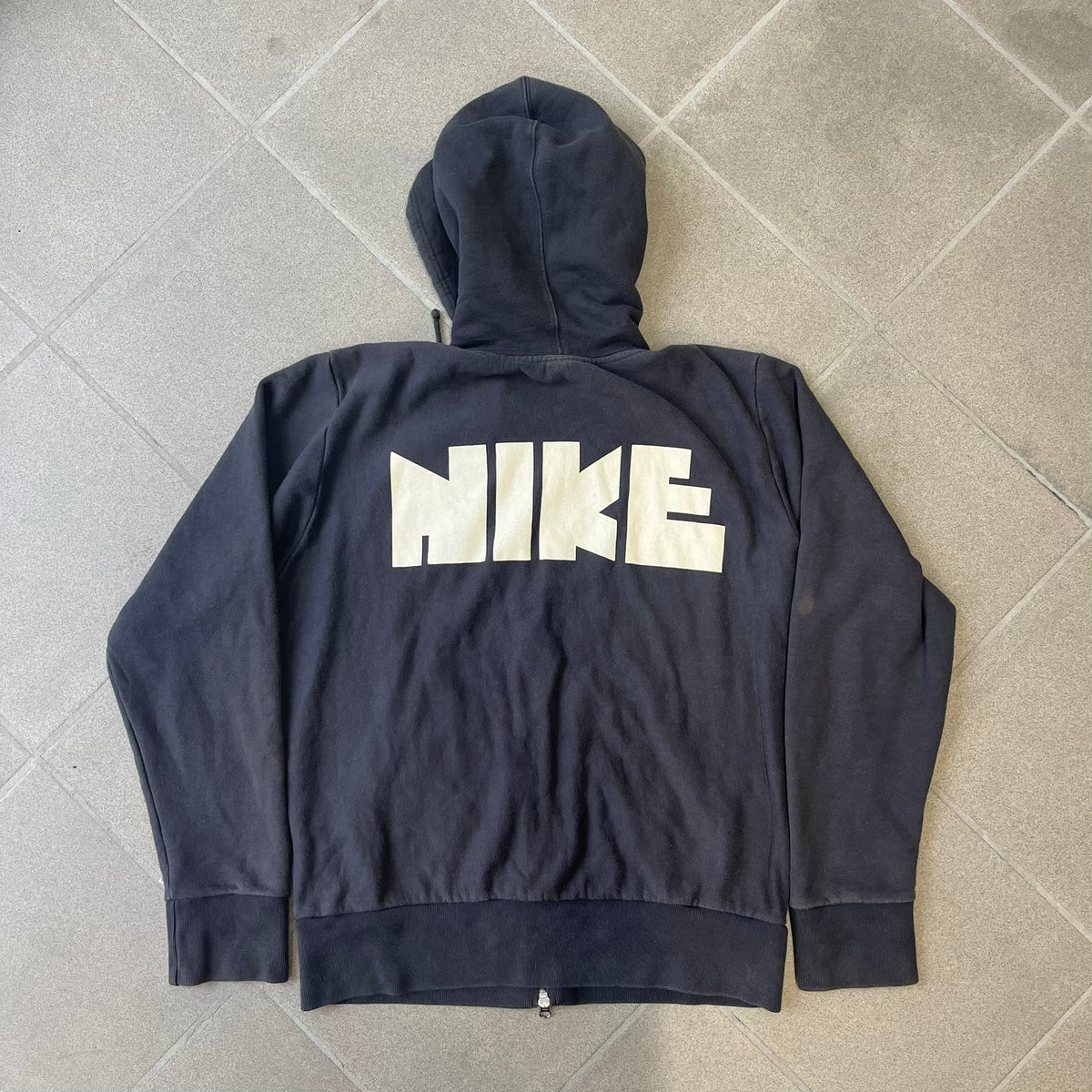 Nike 2010’s Big Logo Black Hoodie is a limited-edition black washed zip hoodie made in Portugal, with a white Nike logo on the chest and a larger one across the upper back, both non-standard designs. It has a standard zipper and a neck tag styled after Nike’s 1980s tags, and the whole piece is finished to look vintage.