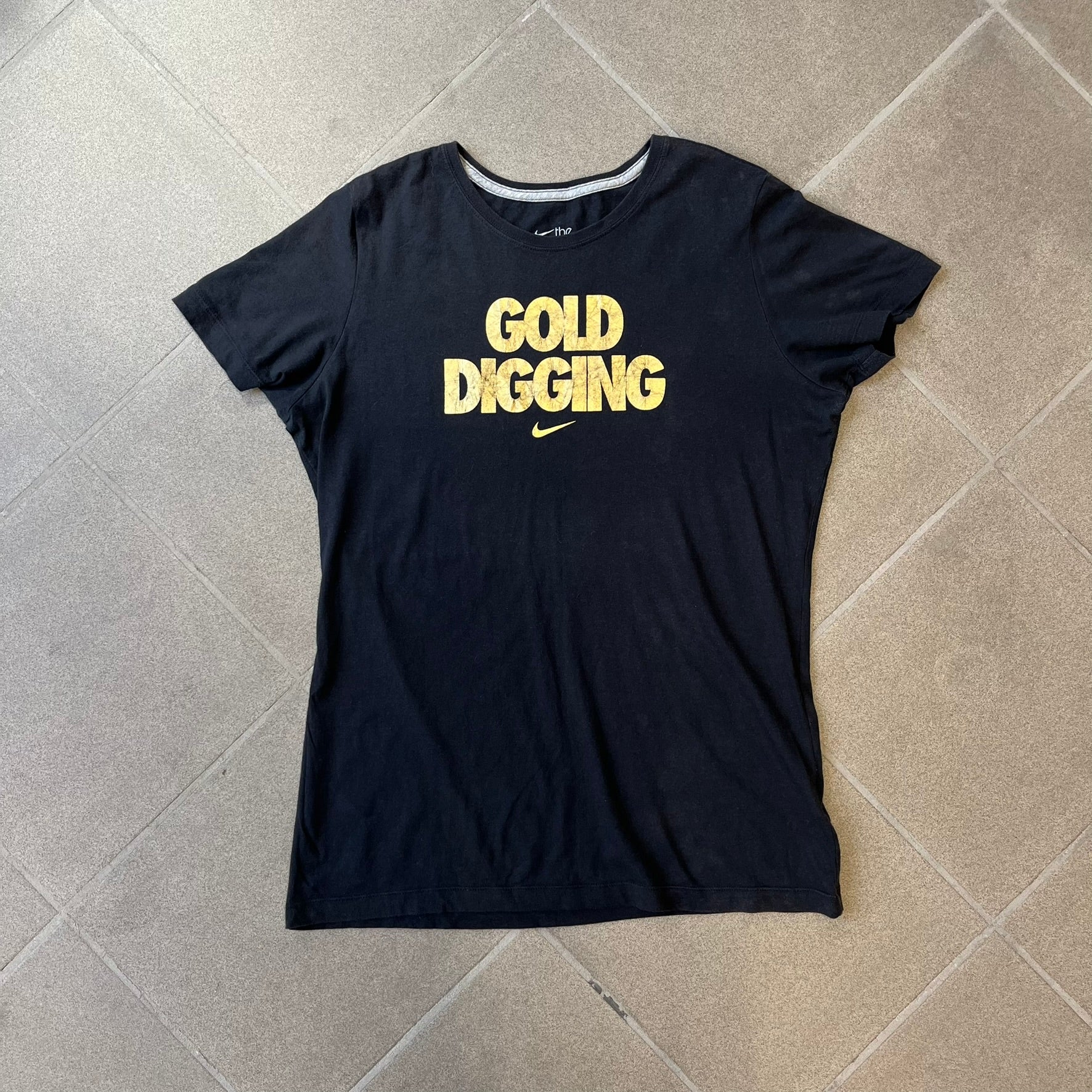 Nike 2012 Olympics “Gold Digging” Black Tee features a black slim-fit T-shirt with gold “Gold Digging” text on the front and a small Nike logo underneath. The design was released in connection with the 2012 Olympics and was later discontinued following controversy surrounding the wording.
