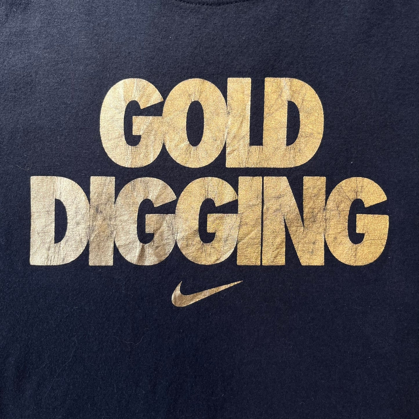 Nike 2012 Olympics “Gold Digging” Black Tee features a black slim-fit T-shirt with gold “Gold Digging” text on the front and a small Nike logo underneath. The design was released in connection with the 2012 Olympics and was later discontinued following controversy surrounding the wording.
