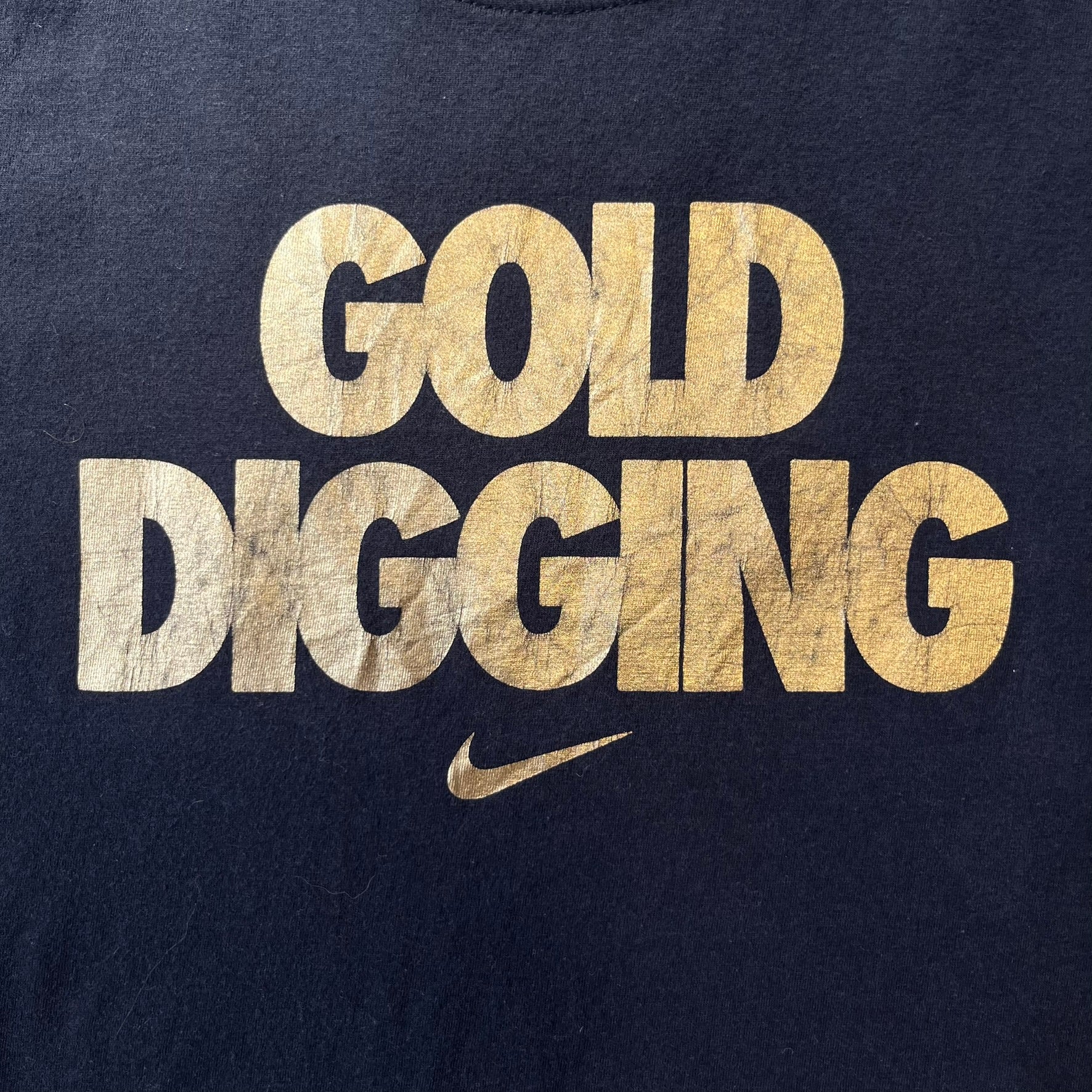 Nike 2012 Olympics “Gold Digging” Black Tee features a black slim-fit T-shirt with gold “Gold Digging” text on the front and a small Nike logo underneath. The design was released in connection with the 2012 Olympics and was later discontinued following controversy surrounding the wording.
