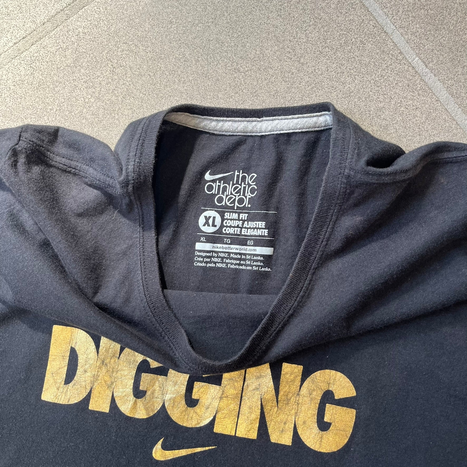 Nike 2012 Olympics “Gold Digging” Black Tee features a black slim-fit T-shirt with gold “Gold Digging” text on the front and a small Nike logo underneath. The design was released in connection with the 2012 Olympics and was later discontinued following controversy surrounding the wording.
