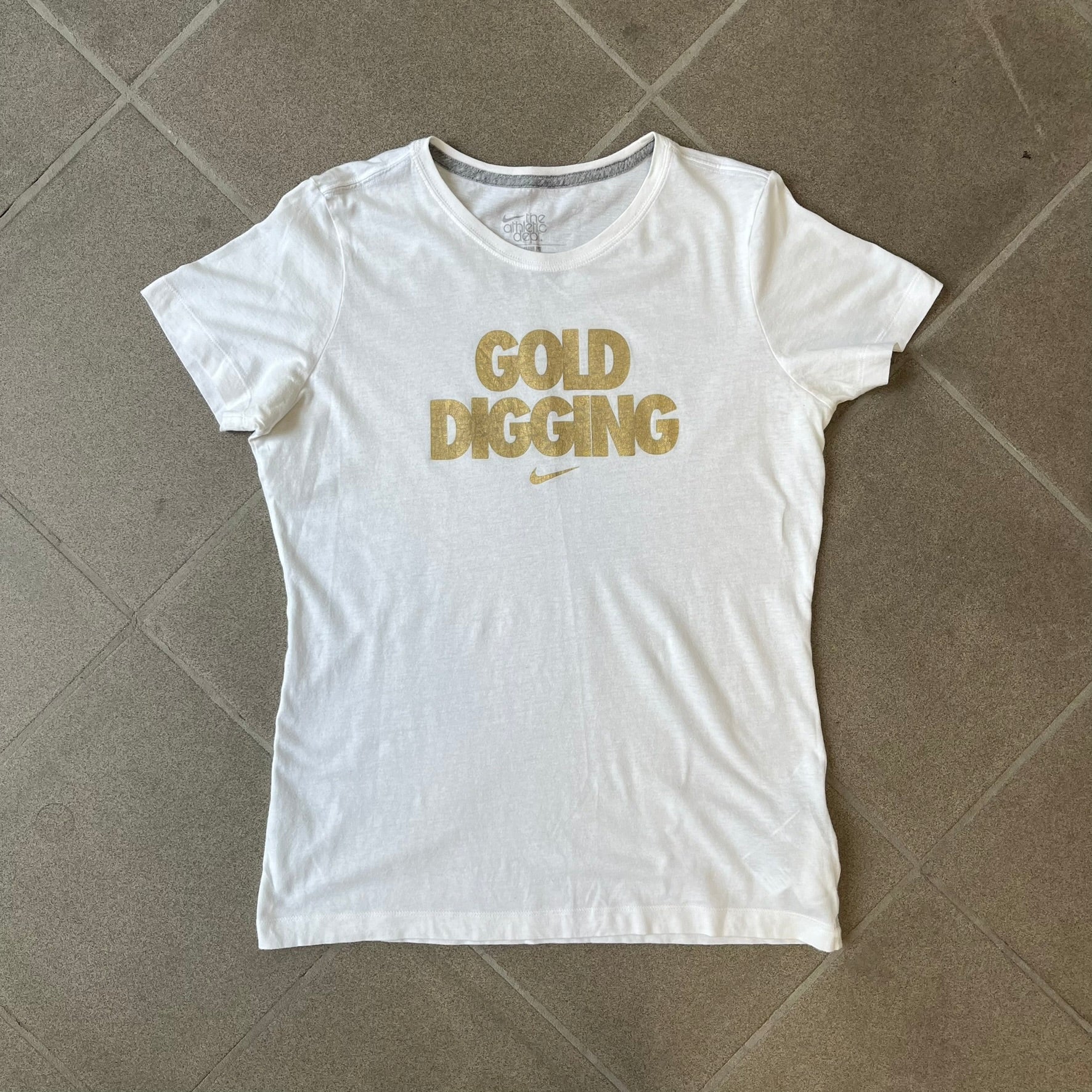 Nike 2012 Olympics “Gold Digging” White Tee features a white slim-fit T-shirt with gold “Gold Digging” text on the front and a small Nike logo underneath. The design was released in connection with the 2012 Olympics and was later discontinued following controversy surrounding the wording.