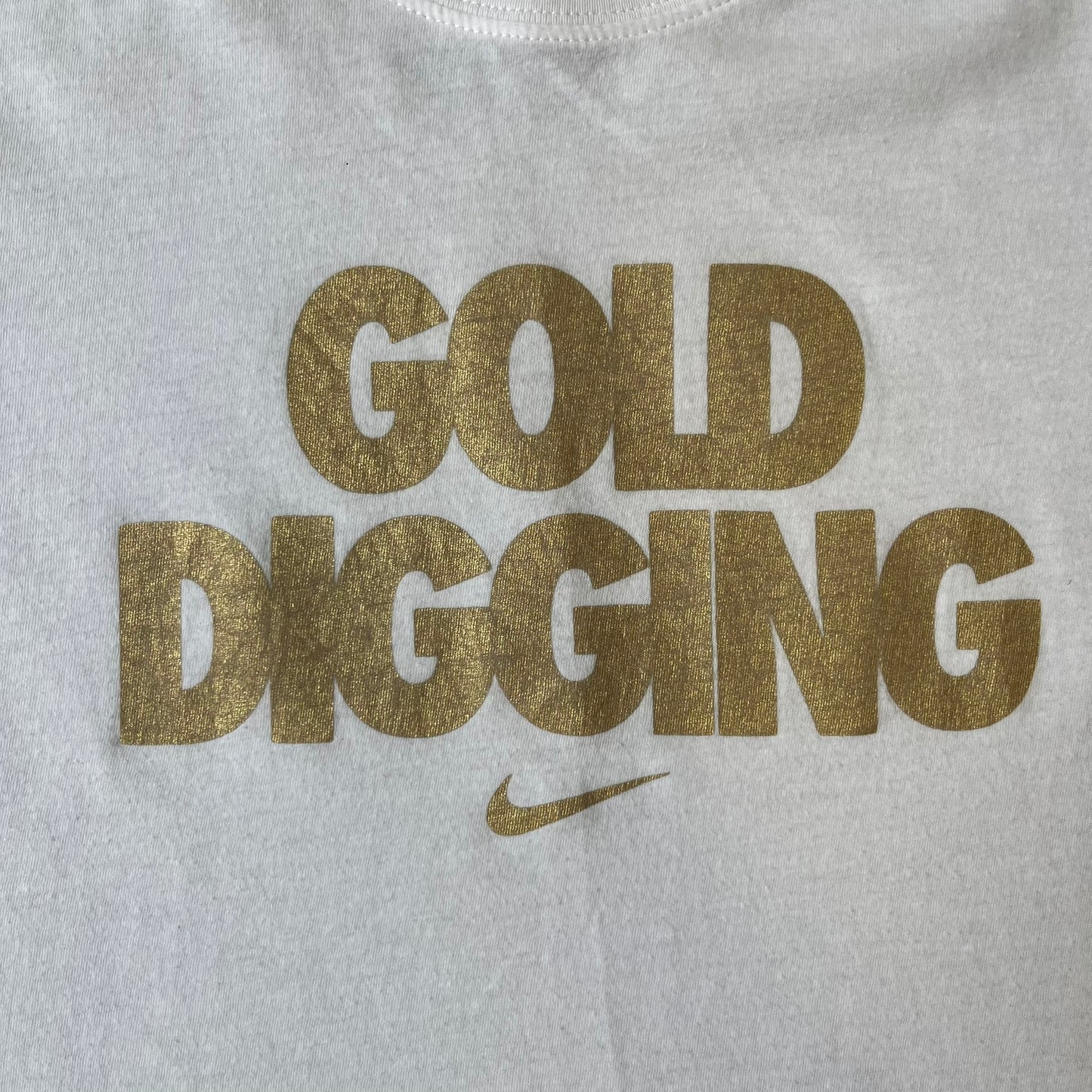 Nike 2012 Olympics “Gold Digging” White Tee features a white slim-fit T-shirt with gold “Gold Digging” text on the front and a small Nike logo underneath. The design was released in connection with the 2012 Olympics and was later discontinued following controversy surrounding the wording.