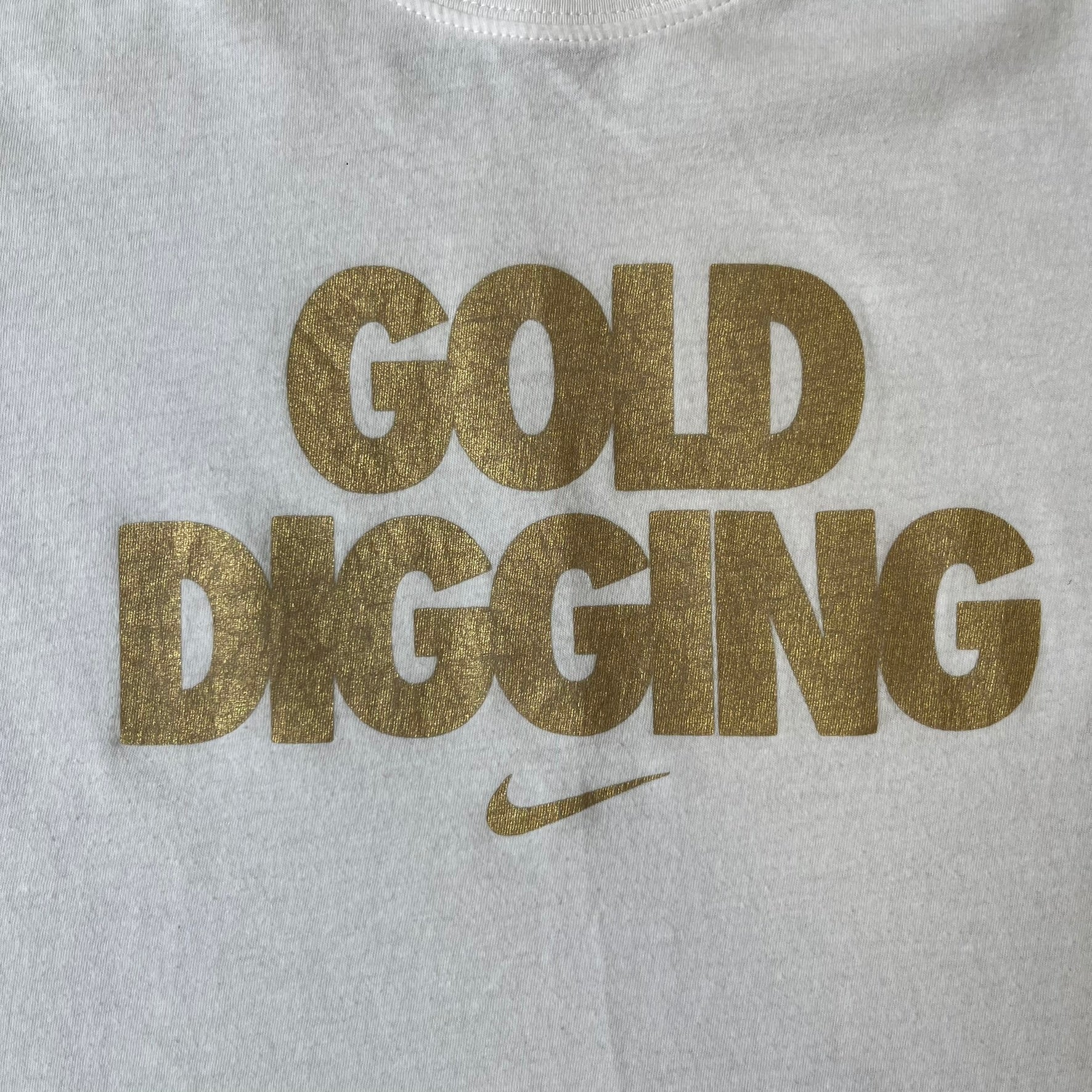 Nike 2012 Olympics “Gold Digging” White Tee features a white slim-fit T-shirt with gold “Gold Digging” text on the front and a small Nike logo underneath. The design was released in connection with the 2012 Olympics and was later discontinued following controversy surrounding the wording.