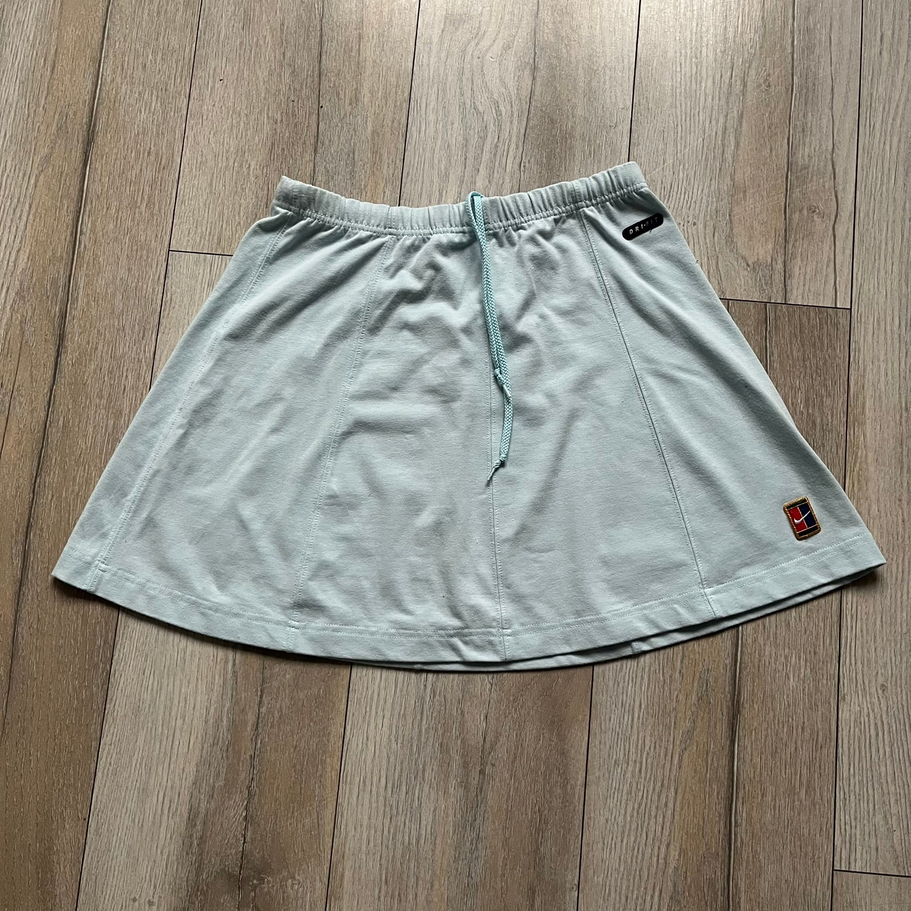 Nike 90’s Challenge Court Tennis Skirt is a baby blue women’s tennis skirt made in Thailand. It features a small green and red Nike Court logo patch at the lower hem, a Dri-FIT patch at the top, and an adjustable waistband with drawstrings.&nbsp;
