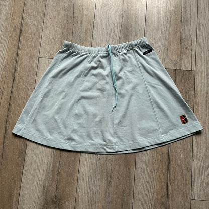Nike 90’s Challenge Court Tennis Skirt is a baby blue women’s tennis skirt made in Thailand. It features a small green and red Nike Court logo patch at the lower hem, a Dri-FIT patch at the top, and an adjustable waistband with drawstrings.&nbsp;