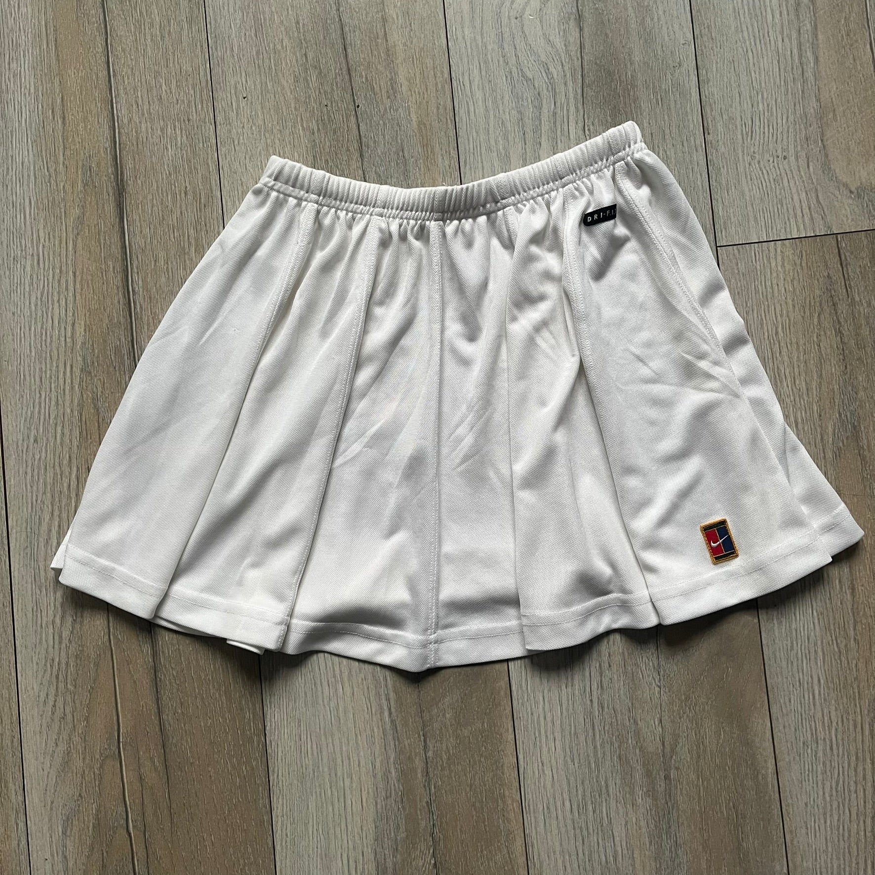 Nike 90’s Challenge Court Tennis Skirt is a white women’s tennis skirt made in USA. It features a small green and red Nike Court logo patch at the lower hem, a Dri-FIT patch at the top, and an adjustable waistband with drawstrings.
