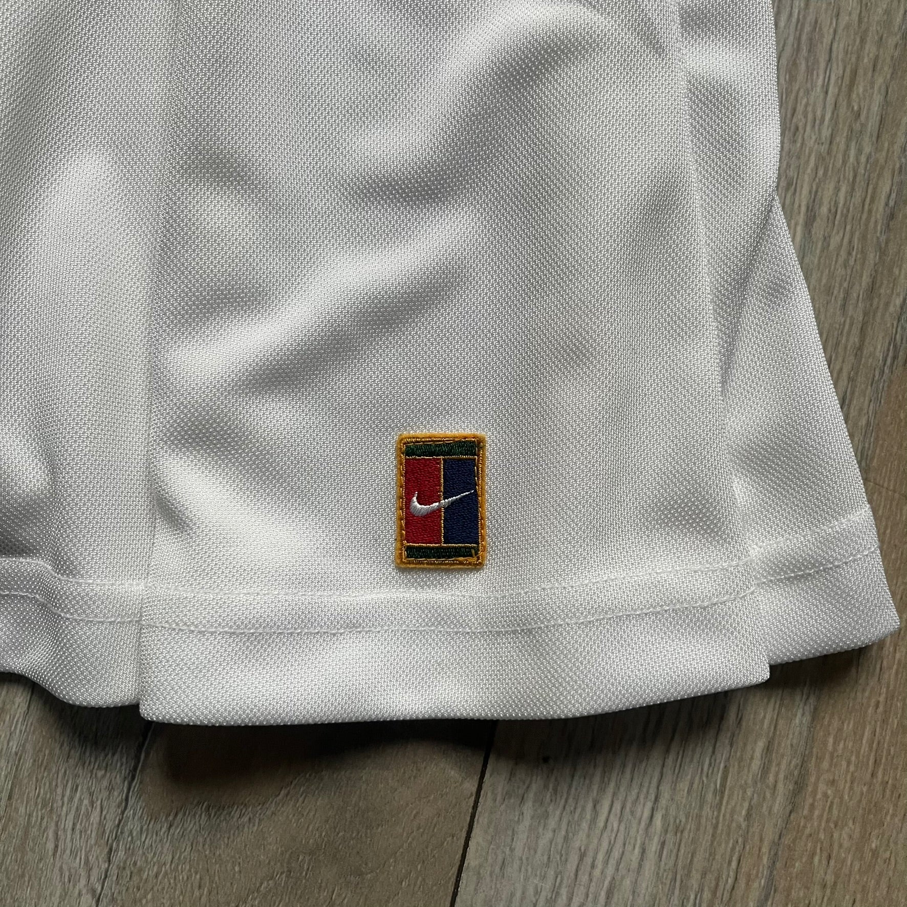 Nike 90’s Challenge Court Tennis Skirt is a white women’s tennis skirt made in USA. It features a small green and red Nike Court logo patch at the lower hem, a Dri-FIT patch at the top, and an adjustable waistband with drawstrings.