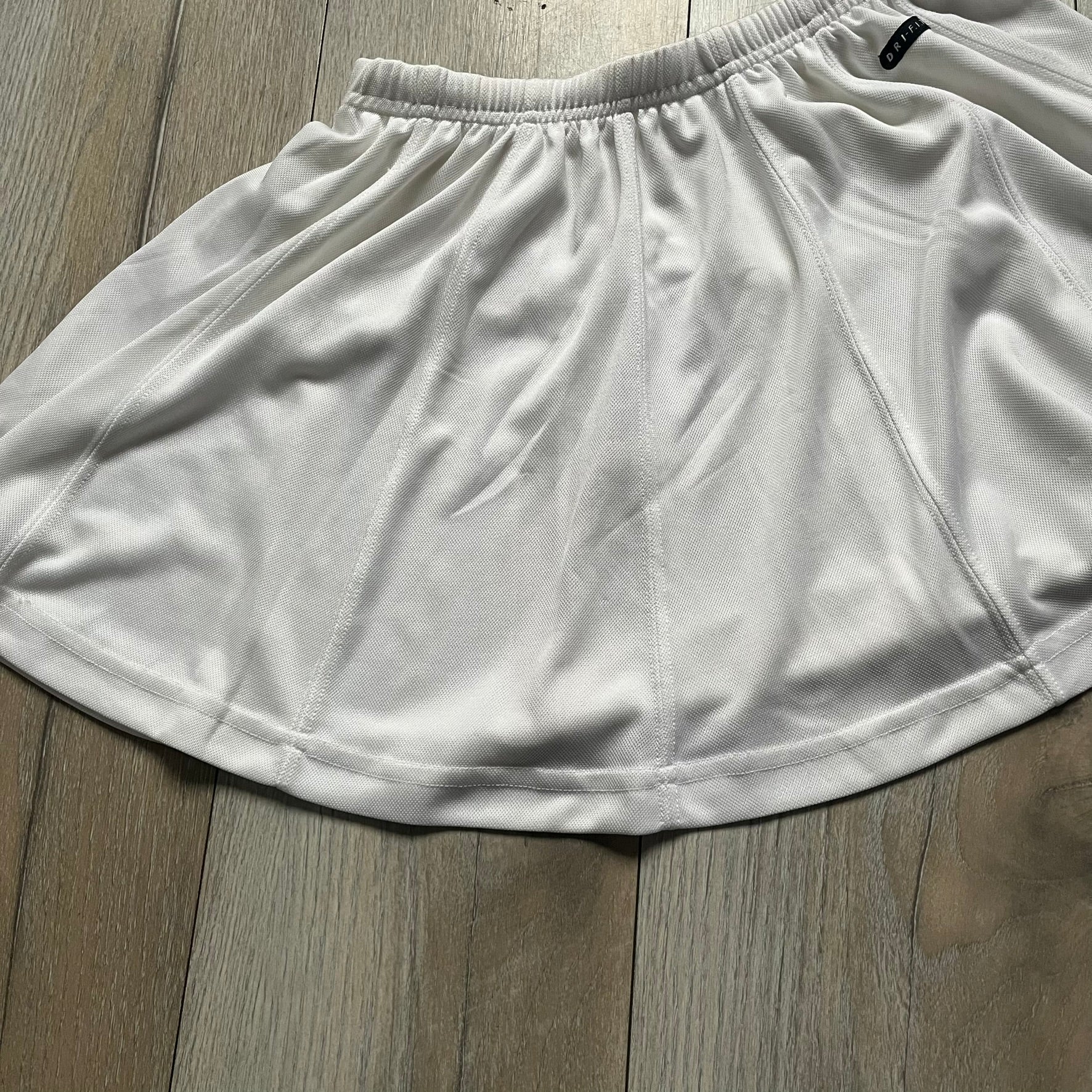 Nike 90’s Challenge Court Tennis Skirt is a white women’s tennis skirt made in USA. It features a small green and red Nike Court logo patch at the lower hem, a Dri-FIT patch at the top, and an adjustable waistband with drawstrings.