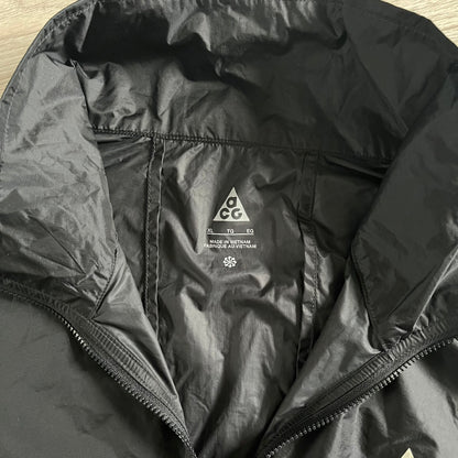 Nike ACG “Cinder Cone” Windbreaker in black, crafted from 100% recycled nylon ripstop. Features a lightweight, water-repellent shell, half-zip closure, and extended collar for added coverage. Designed with no shoulder seams for mobility and finished with printed ACG chest branding, two zippered pockets, and an internal baby carabiner.