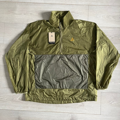 Olive Nike ACG Cinder Cone 1/2 Zip Jacket made from durable nylon ripstop with water-repellent finish. Designed with no shoulder seams for mobility, elastic cuffs, reinforced hem, and two zip pockets. Includes printed chest logo and internal ACG carabiner, with packable oversized construction.