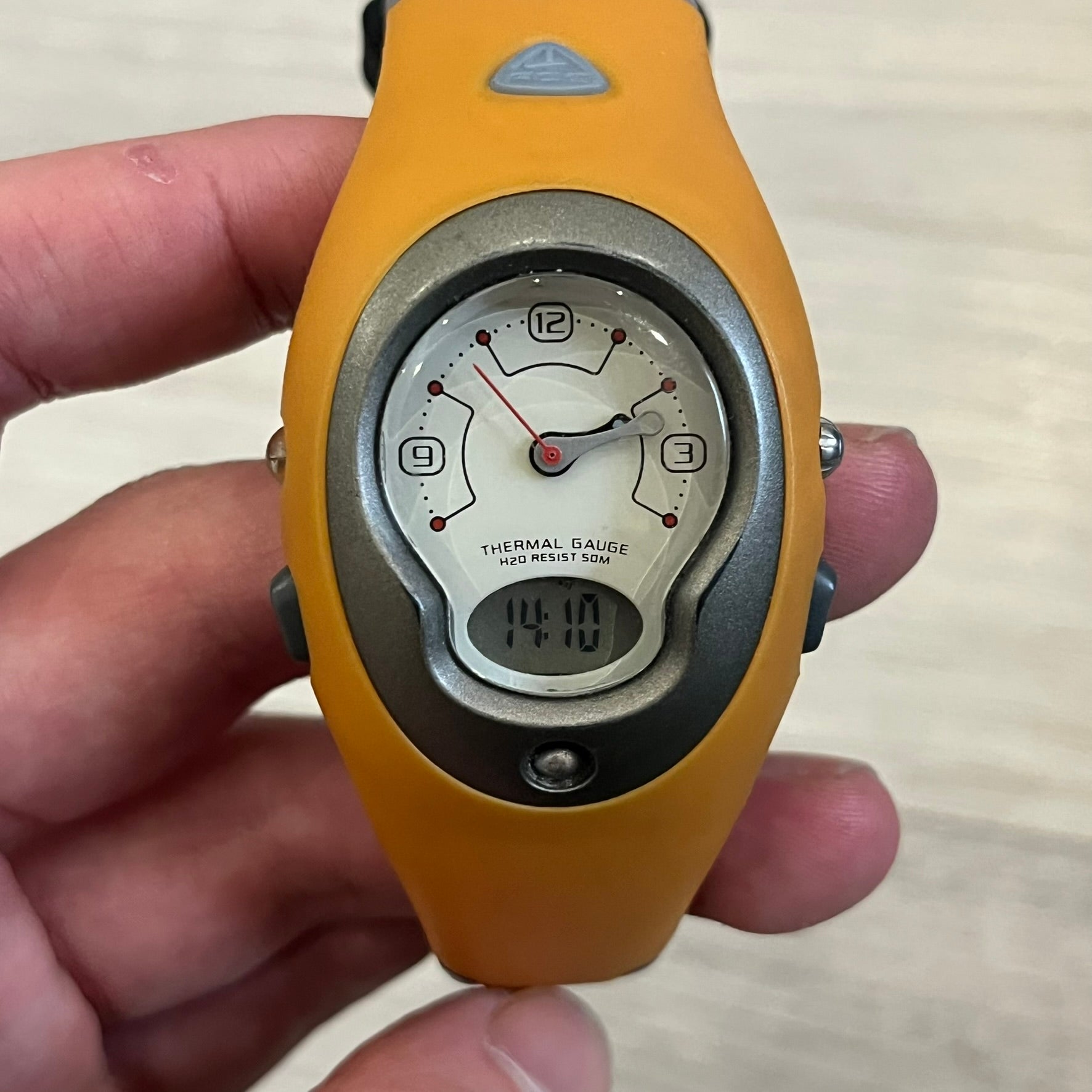 Nike ACG Thermal Gauge Watch featuring an ani-digi display with temperature, date, timer, alarm, and counter functions. Designed with a durable stainless-steel and plastic build, a white dial with grey outline, and an orange strap with the ACG logo. Water-resistant up to 50m, this Nike ACG outdoor watch blends utility with signature 90s–2000s adventure styling.