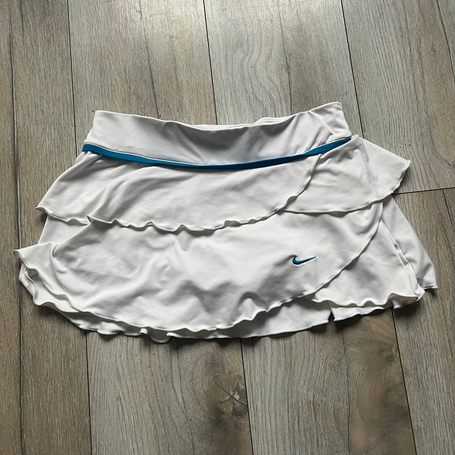 Nike Active Frilly 00’s Tennis Skirt is a women’s tennis skort with integrated inner shorts for support. The white skirt features multiple irregular frilly layers, creating a layered look, with a blue embroidered Nike Swoosh on the front layer. A blue contrast line separates the waistband from the skirt, emphasizing its sporty 2000s design.