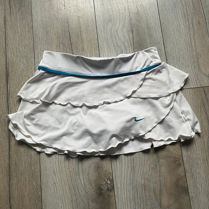 Nike Active Frilly 00’s Tennis Skirt is a women’s tennis skort with integrated inner shorts for support. The white skirt features multiple irregular frilly layers, creating a layered look, with a blue embroidered Nike Swoosh on the front layer. A blue contrast line separates the waistband from the skirt, emphasizing its sporty 2000s design.