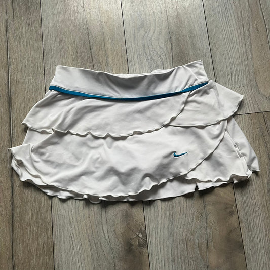 Nike Active Frilly 00’s Tennis Skirt is a women’s tennis skort with integrated inner shorts for support. The white skirt features multiple irregular frilly layers, creating a layered look, with a blue embroidered Nike Swoosh on the front layer. A blue contrast line separates the waistband from the skirt, emphasizing its sporty 2000s design.