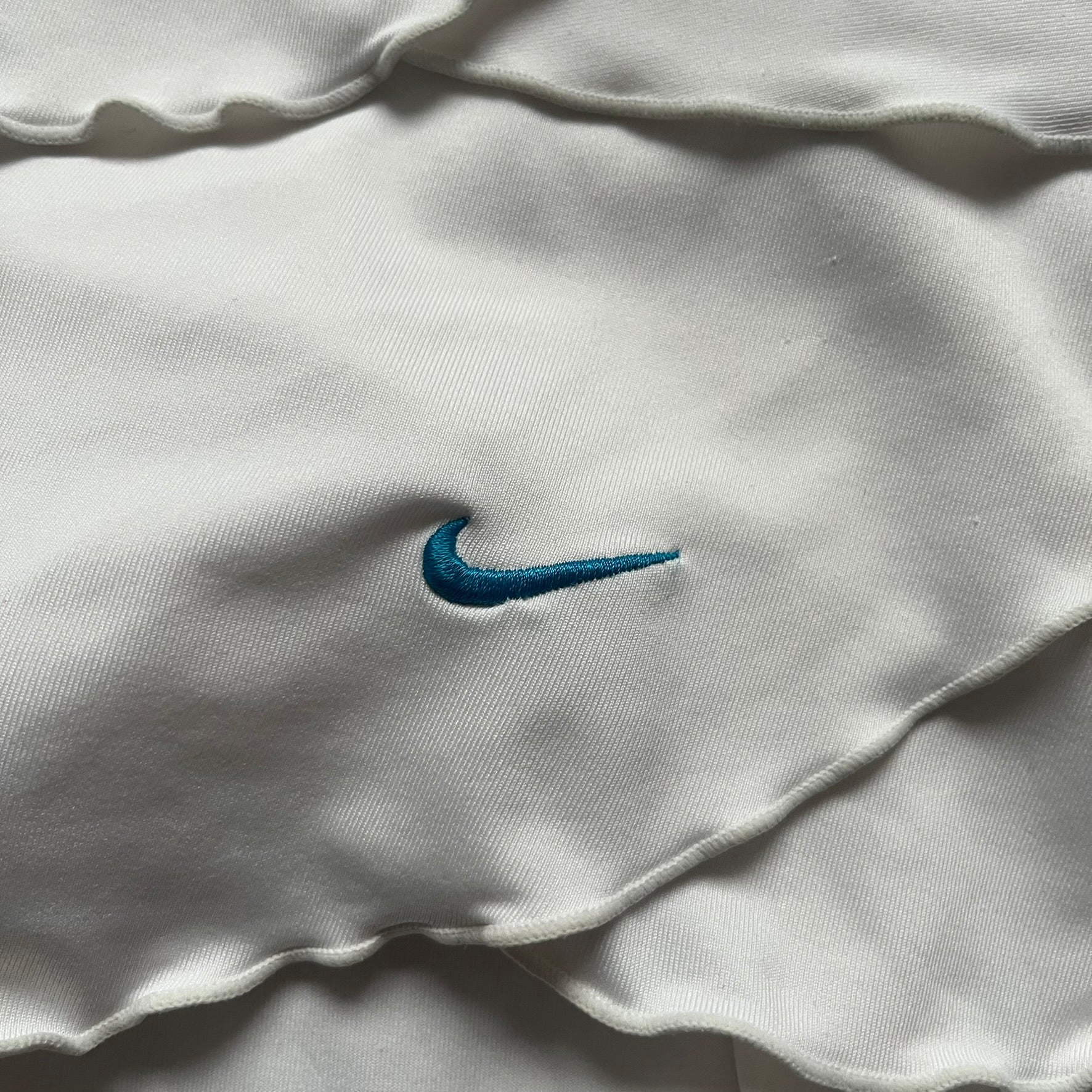 Nike Active Frilly 00’s Tennis Skirt is a women’s tennis skort with integrated inner shorts for support. The white skirt features multiple irregular frilly layers, creating a layered look, with a blue embroidered Nike Swoosh on the front layer. A blue contrast line separates the waistband from the skirt, emphasizing its sporty 2000s design.