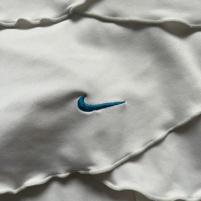 Nike Active Frilly 00’s Tennis Skirt is a women’s tennis skort with integrated inner shorts for support. The white skirt features multiple irregular frilly layers, creating a layered look, with a blue embroidered Nike Swoosh on the front layer. A blue contrast line separates the waistband from the skirt, emphasizing its sporty 2000s design.
