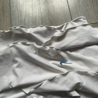 Nike Active Frilly 00’s Tennis Skirt is a women’s tennis skort with integrated inner shorts for support. The white skirt features multiple irregular frilly layers, creating a layered look, with a blue embroidered Nike Swoosh on the front layer. A blue contrast line separates the waistband from the skirt, emphasizing its sporty 2000s design.