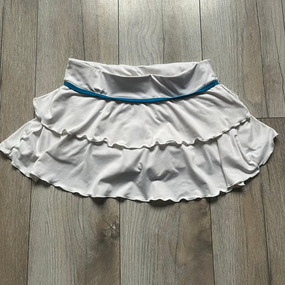 Nike Active Frilly 00’s Tennis Skirt is a women’s tennis skort with integrated inner shorts for support. The white skirt features multiple irregular frilly layers, creating a layered look, with a blue embroidered Nike Swoosh on the front layer. A blue contrast line separates the waistband from the skirt, emphasizing its sporty 2000s design.