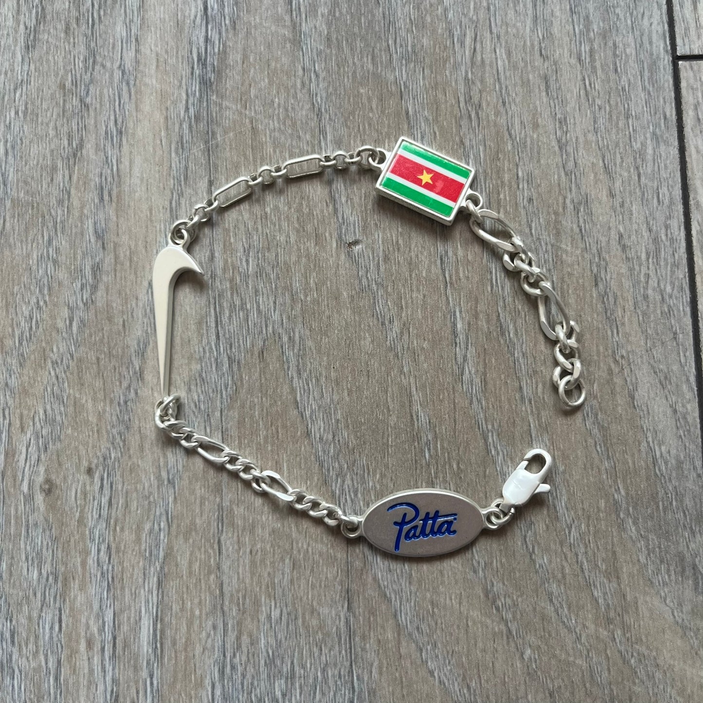 Nike Air Max x Patta Bracelet is a metal chain bracelet released as part of the Nike x Patta Air Max 1 collaboration. The bracelet features linked charms including Nike, Patta, and Suriname flag motifs, all connected along a light silver-tone chain. It was included exclusively with special box editions of the Patta x Nike Air Max 1 release and comes with a black Nike/Patta dust bag.
