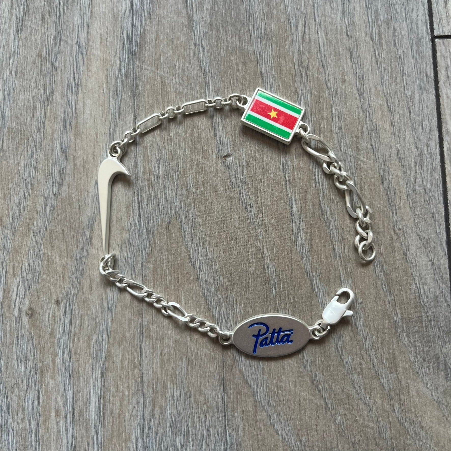Nike Air Max x Patta Bracelet is a metal chain bracelet released as part of the Nike x Patta Air Max 1 collaboration. The bracelet features linked charms including Nike, Patta, and Suriname flag motifs, all connected along a light silver-tone chain. It was included exclusively with special box editions of the Patta x Nike Air Max 1 release and comes with a black Nike/Patta dust bag.