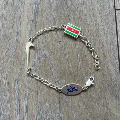 Nike Air Max x Patta Bracelet is a metal chain bracelet released as part of the Nike x Patta Air Max 1 collaboration. The bracelet features linked charms including Nike, Patta, and Suriname flag motifs, all connected along a light silver-tone chain. It was included exclusively with special box editions of the Patta x Nike Air Max 1 release and comes with a black Nike/Patta dust bag.