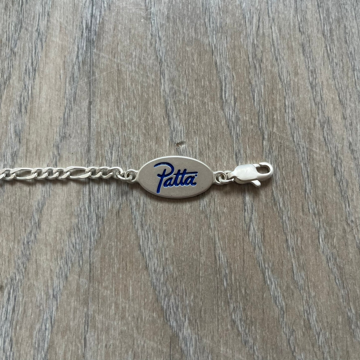 Nike Air Max x Patta Bracelet is a metal chain bracelet released as part of the Nike x Patta Air Max 1 collaboration. The bracelet features linked charms including Nike, Patta, and Suriname flag motifs, all connected along a light silver-tone chain. It was included exclusively with special box editions of the Patta x Nike Air Max 1 release and comes with a black Nike/Patta dust bag.