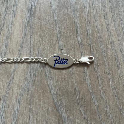 Nike Air Max x Patta Bracelet is a metal chain bracelet released as part of the Nike x Patta Air Max 1 collaboration. The bracelet features linked charms including Nike, Patta, and Suriname flag motifs, all connected along a light silver-tone chain. It was included exclusively with special box editions of the Patta x Nike Air Max 1 release and comes with a black Nike/Patta dust bag.