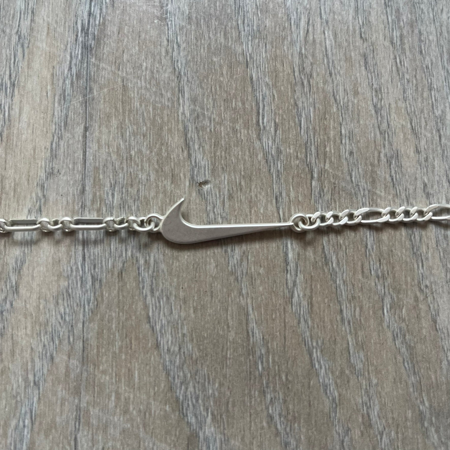 Nike Air Max x Patta Bracelet is a metal chain bracelet released as part of the Nike x Patta Air Max 1 collaboration. The bracelet features linked charms including Nike, Patta, and Suriname flag motifs, all connected along a light silver-tone chain. It was included exclusively with special box editions of the Patta x Nike Air Max 1 release and comes with a black Nike/Patta dust bag.