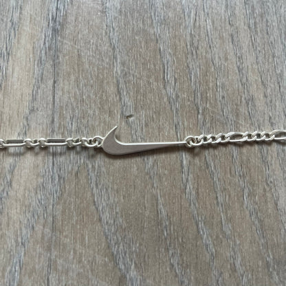 Nike Air Max x Patta Bracelet is a metal chain bracelet released as part of the Nike x Patta Air Max 1 collaboration. The bracelet features linked charms including Nike, Patta, and Suriname flag motifs, all connected along a light silver-tone chain. It was included exclusively with special box editions of the Patta x Nike Air Max 1 release and comes with a black Nike/Patta dust bag.