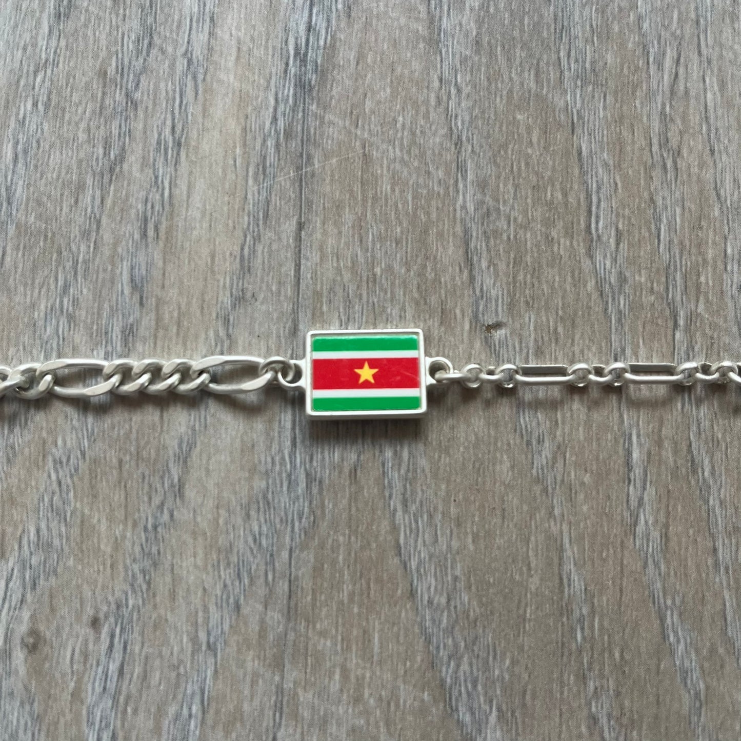 Nike Air Max x Patta Bracelet is a metal chain bracelet released as part of the Nike x Patta Air Max 1 collaboration. The bracelet features linked charms including Nike, Patta, and Suriname flag motifs, all connected along a light silver-tone chain. It was included exclusively with special box editions of the Patta x Nike Air Max 1 release and comes with a black Nike/Patta dust bag.