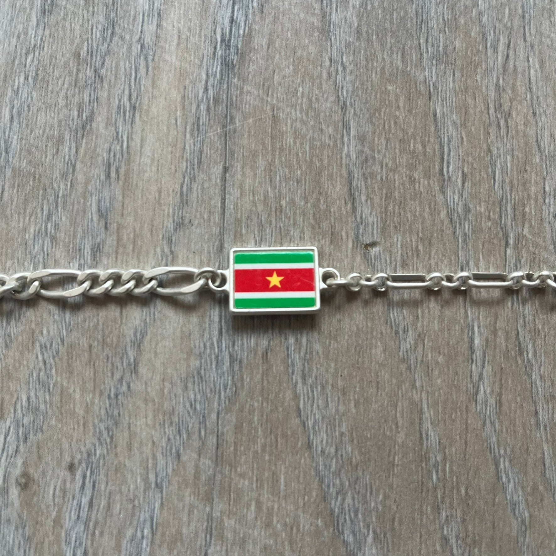 Nike Air Max x Patta Bracelet is a metal chain bracelet released as part of the Nike x Patta Air Max 1 collaboration. The bracelet features linked charms including Nike, Patta, and Suriname flag motifs, all connected along a light silver-tone chain. It was included exclusively with special box editions of the Patta x Nike Air Max 1 release and comes with a black Nike/Patta dust bag.