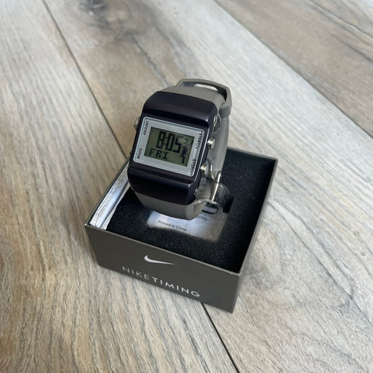 Nike Anvil Comold Reg Digital Watch WC0038 is a digital wristwatch featuring a dark purple/violet aluminum and stainless steel case with a white-outlined display. The watch includes four side buttons, a digital screen, and grey rubber straps, constructed from a mix of aluminum, stainless steel, and plastic. It is water resistant up to 100 meters, powered by fresh batteries, and comes with the original box.